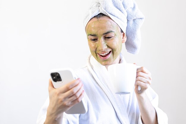 Young woman with a green beauty mask on her face with a cup and a smartphone