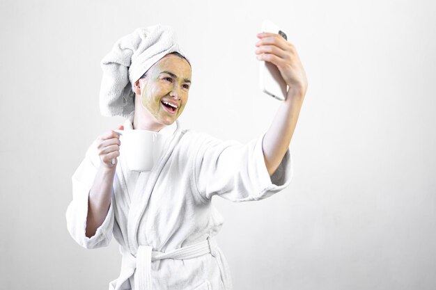 Young woman with a green beauty mask on her face with a cup and a smartphone