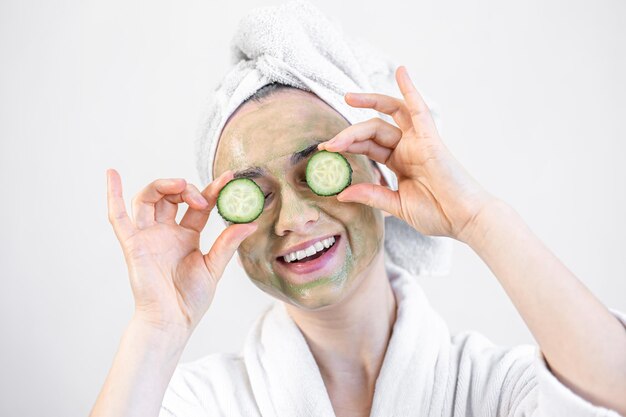 Young woman with a green face mask and fresh cucumbers in a white bathrobe