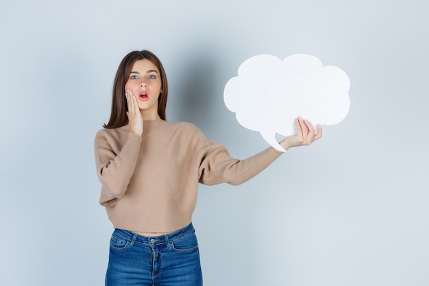 Young woman with hand on cheek, keeping paper speech bubble in sweater, jeans and looking surprised , front view.