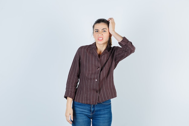 Young woman with hand on head, grimacing in striped shirt, jeans and looking agitated. front view.