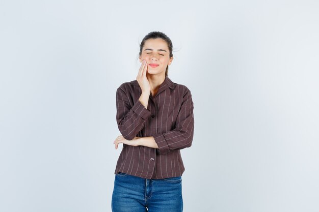 Young woman with hand near mouth, having toothache in striped shirt, jeans and looking exhausted. front view.