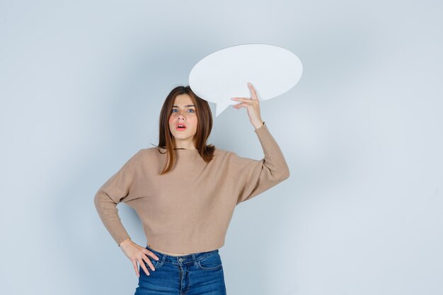 Young woman with hand on waist, raising paper speech bubble in sweater, jeans and looking surprised , front view.