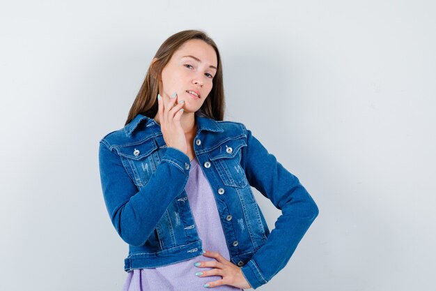 Young woman with hand on waist, touching her jaw in t-shirt, jacket and looking delicate , front view.