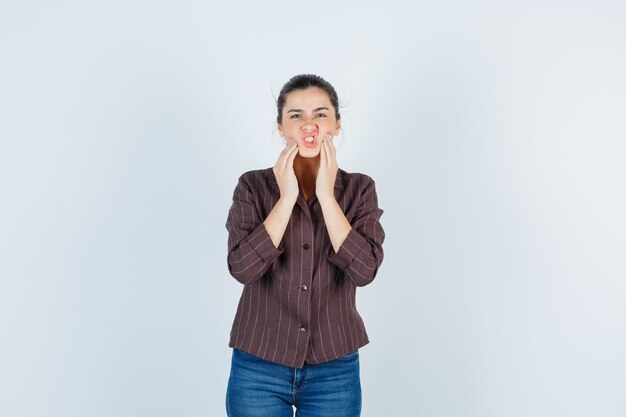 Young woman with hands on cheeks, keeping mouth open, grimacing in striped shirt, jeans and looking cute , front view.