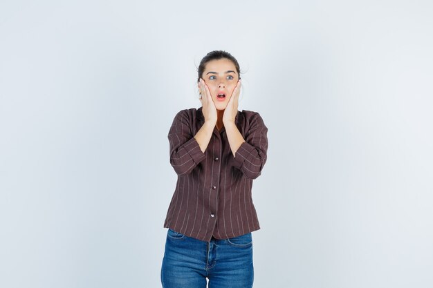 Молодая женщина с руками на щеках, широко открытым ртом в полосатой рубашке, джинсах и шокированным видом, вид спереди.