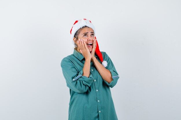 Молодая женщина с руками на щеках, открывая рот в рубашке, шляпе Санта-Клауса и выглядя в ужасе. передний план.