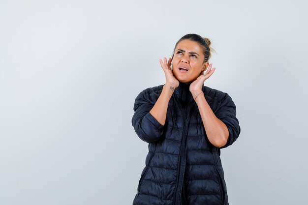 Young woman with hands near ears, looking up in puffer jacket and looking confused. front view.