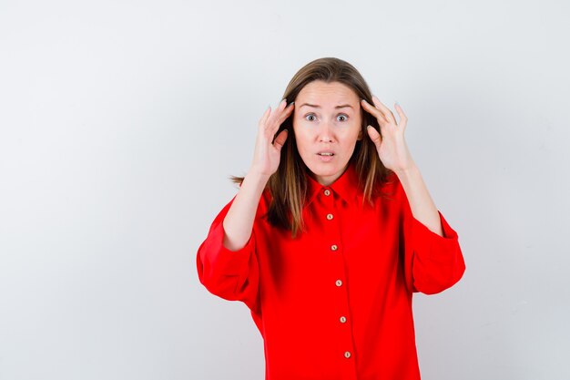 Young woman with hands near ears in red blouse and looking puzzled. front view.