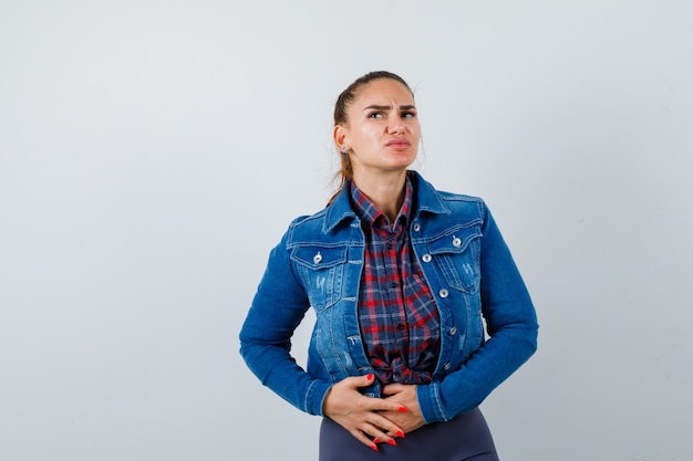 Giovane donna con le mani sullo stomaco in camicia a scacchi, giacca di jeans e sguardo doloroso, vista frontale.