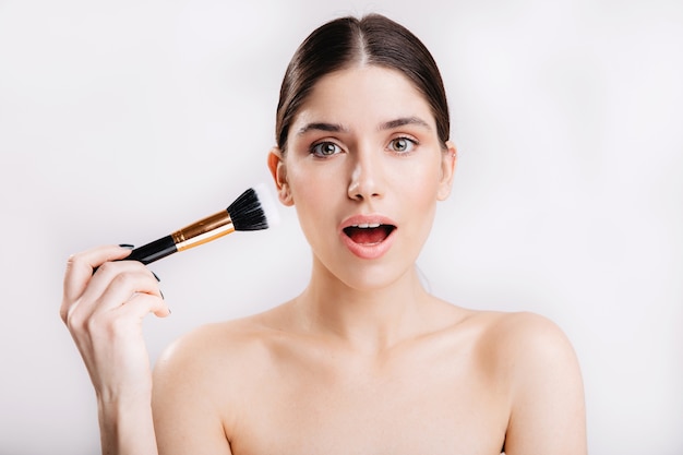 Young woman with healthy skin posing in surprise with brush for foundation on white wall.