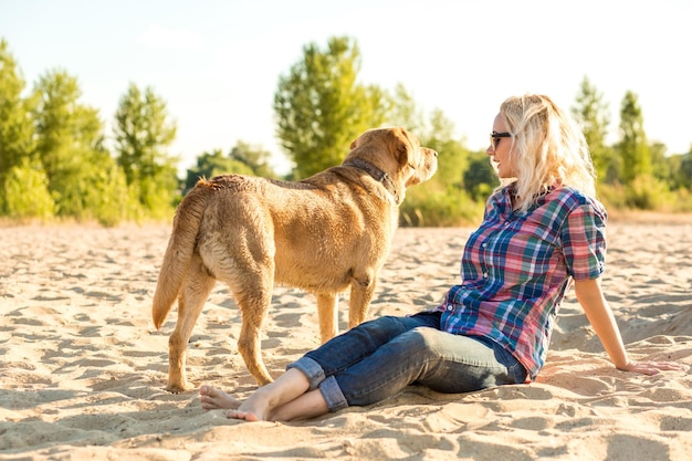 해변에서 그녀의 강아지와 함께 젊은 여자. 한 젊은 여성이 강아지와 함께 모래 위에 앉아 있습니다. 래브라도