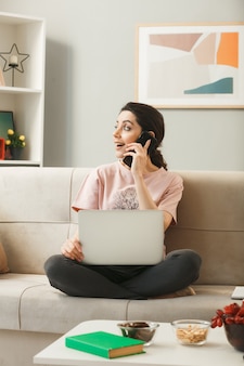 ノートパソコンを持つ若い女性は、リビングルームのコーヒーテーブルの後ろのソファに座って電話で話します