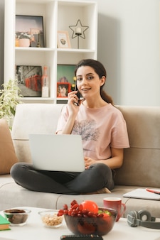 Giovane donna con laptop parla al telefono seduta sul divano dietro il tavolino nel soggiorno