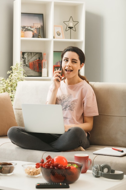 ノートパソコンを持つ若い女性は、リビングルームのコーヒーテーブルの後ろのソファに座って電話で話します