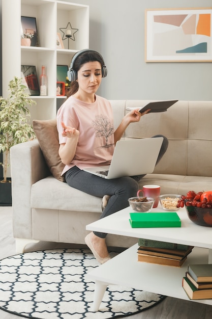 Young woman with laptop wearing headphones holding notebook with pen sitting on sofa behind coffee table in living room