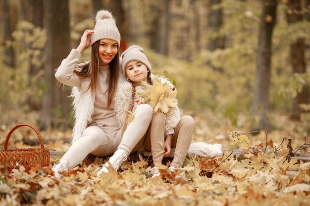 Young woman with little girl sitting on a fallen tree trunk in autumn forest. Brunette woman play with her daughter. Girl wearing beige sweater and mother wearing white clothes.