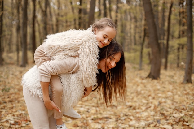 Young woman with little girl standing in autumn forest. Brunette woman holding her daughter piggyback. Girl wearing beige sweater and mother wearing white clothes.