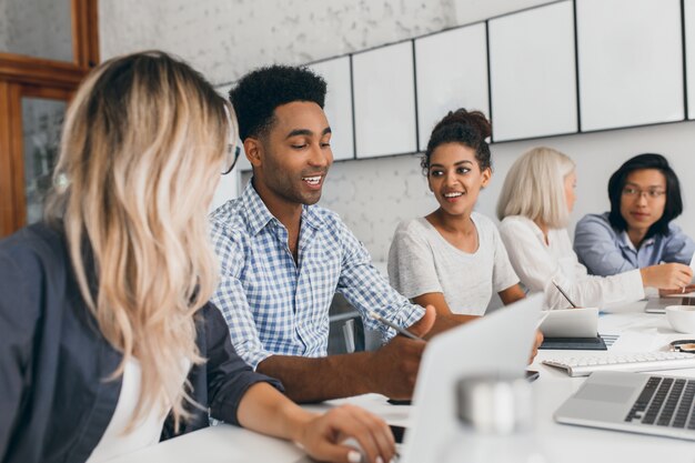 Young woman with long blonde hair listening african man in blue shirt which using laptop. Indoor portrait of black and asian office workers talking during conference.