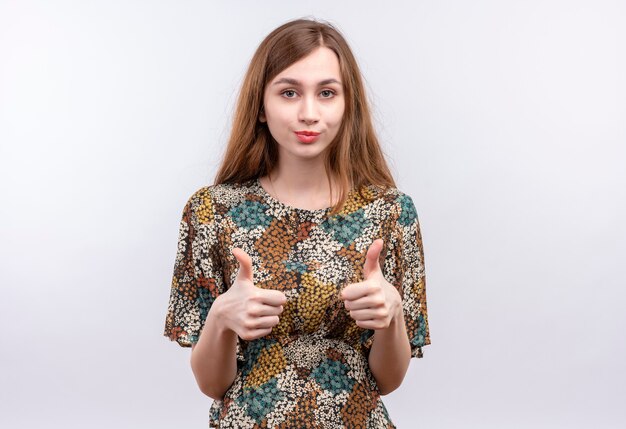 Молодая женщина с длинными волосами, одетая в красочное платье, уверенно улыбается, показывает палец вверх обеими руками, стоя над белой стеной