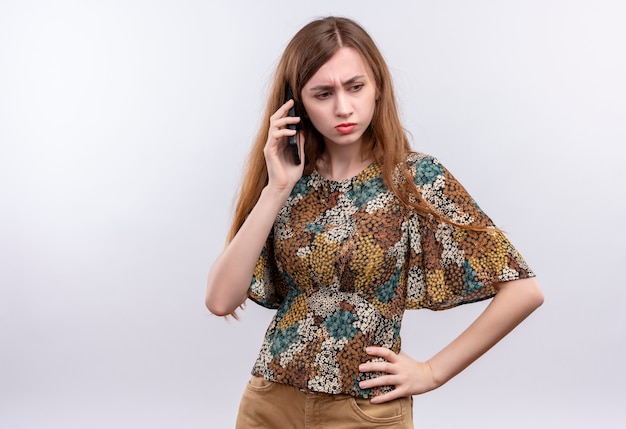 Young woman with long hair wearing colorful dress talking on mobile phone with frowning face standing over white wall