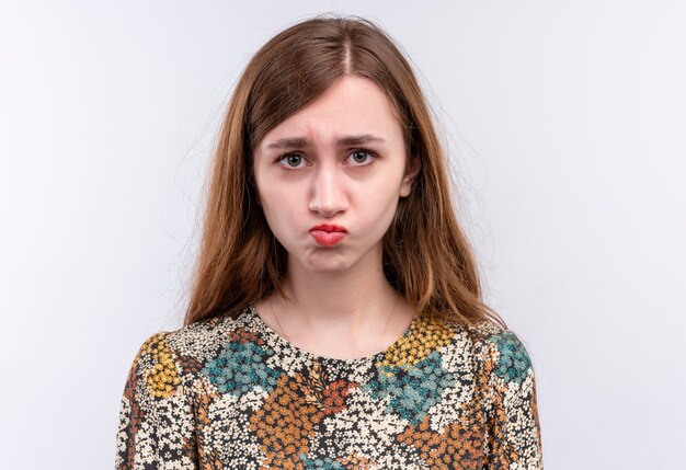 Young woman with long hair wearing colorful dress very  looking with sad expression on face  frowning standing over white wall