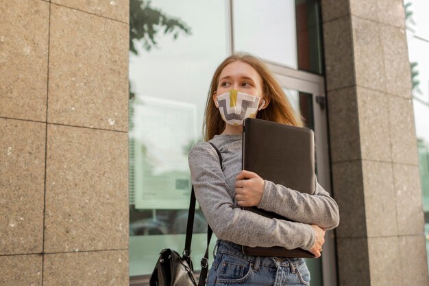 Молодая женщина с медицинской маской держит свой ноутбук на улице