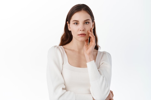 Young woman with nude natural make up, touching hydrated clean facial skin, holding hand on cheek, gazing at front thoughtful, white wall