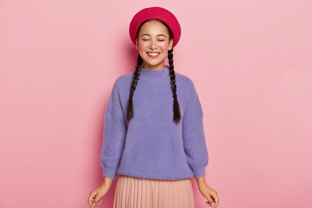 Молодая женщина с радостным выражением лица, держит глаза закрытыми, носит красный берет, теплый фиолетовый свитер и плетеную юбку, получает удовольствие от комплимента, позирует над розовой стеной