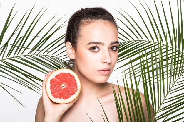 Young woman with perfect skin holding citrus fruit in hand surrounded by palms leaves isolated on white wall