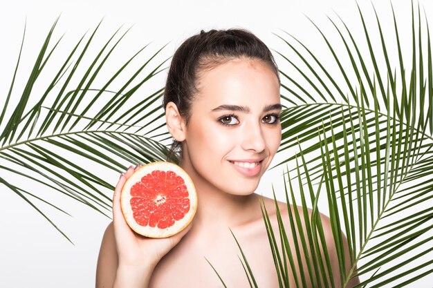 Young woman with perfect skin holding citrus fruit in hand surrounded by palms leaves isolated on white wall