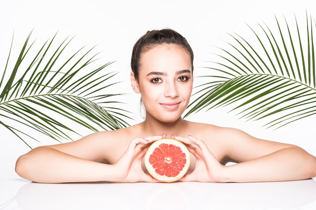 Young woman with perfect skin holding citrus fruit in hands surrounded by palms leaves isolated on white wall