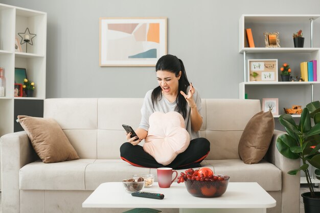 Young woman with pillow holding and looking at phone sitting on sofa behind coffee table in living room