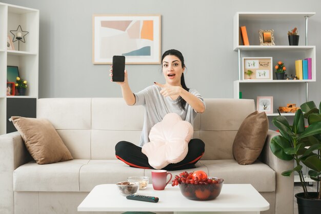 Young woman with pillow holding and points at phone sitting on sofa behind coffee table in living room