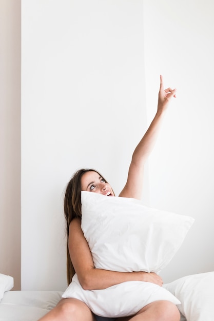 Young woman with pillow sitting on bed pointing upwards