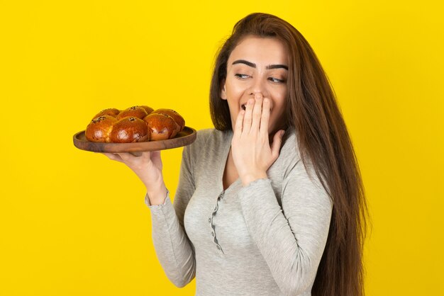 노란색 벽 위에 서 있는 신선한 과자 한 접시를 가진 젊은 여자.