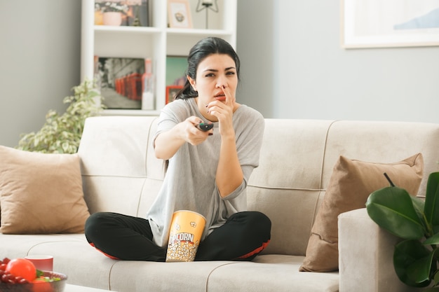Young woman with popcorn bucket holding out tv remote to camera sitting on sofa behind coffee table in living room