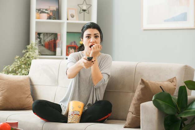 Молодая женщина с ведром для попкорна, протягивая пульт от телевизора к камере, сидит на диване за журнальным столиком в гостиной