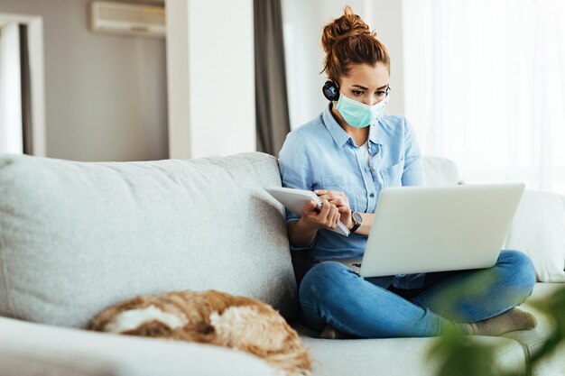 Young woman with protective mask using laptop and writing notes while following classes online from home