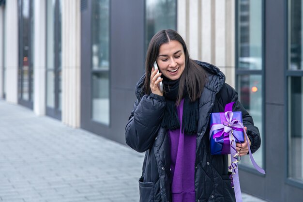 A young woman with a purple gift in her hands talking on the phone outside