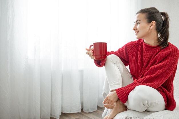 A young woman with a red cup of hot drink in a cozy red sweater is resting on the couch at home copy space.