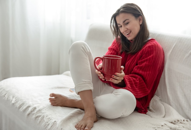 아늑한 빨간 스웨터에 뜨거운 음료의 빨간 컵을 가진 젊은 여성이 가정 복사 공간에서 소파에 쉬고 있습니다.