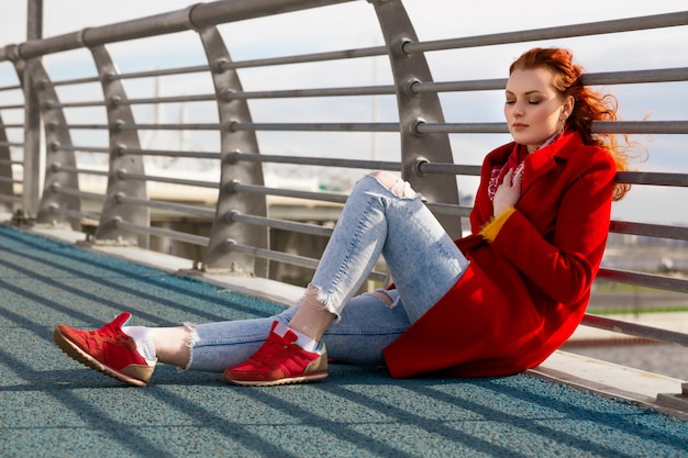 A young woman with red hair in a red coat and red sneakers is sitting on a bridge on a Sunny