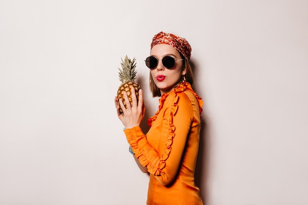 Young woman with red lips is holding pineapple. Portrait of naughty girl in orange outfit and sunglasses on white space.