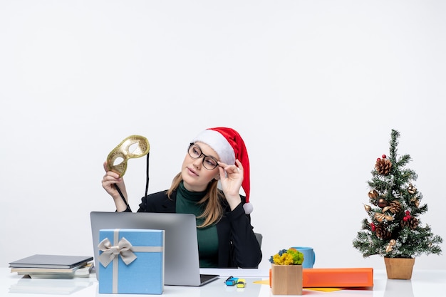 Young woman with santa claus hat eyeglasses and mask sitting at a table with a xmas tree and a gift