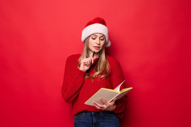 Young woman with Santa hat holds a notepad in her hands and bites a pen checking the list of gifts or children with good behavior isolated on a red wall