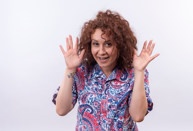 Young woman with short curly hair  in colorful shirt having fun smiling cheerfully holding palms near face standing over white wall