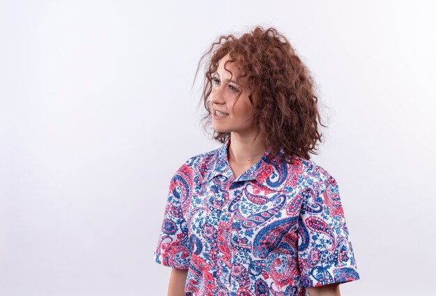 Young woman with short curly hair  in colorful shirt looking aside smiling standing over white wall