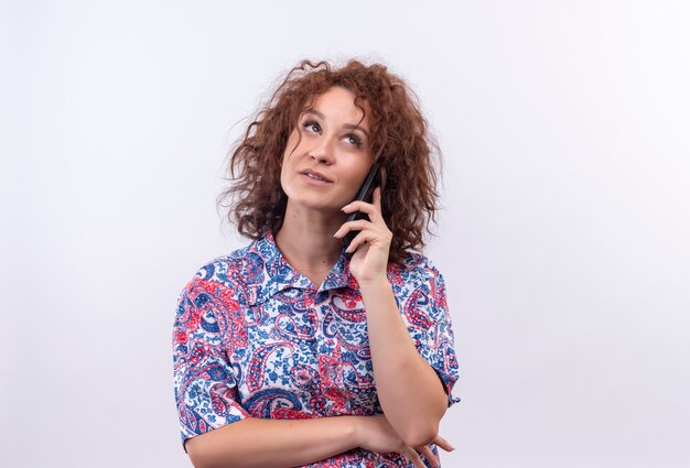 Young woman with short curly hair  in colorful shirt  looking up thinking while talking on mobile phone over white wall