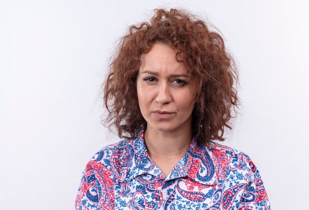 Free photo young woman with short curly hair  in colorful shirt  looking with frowning face standing over white wall